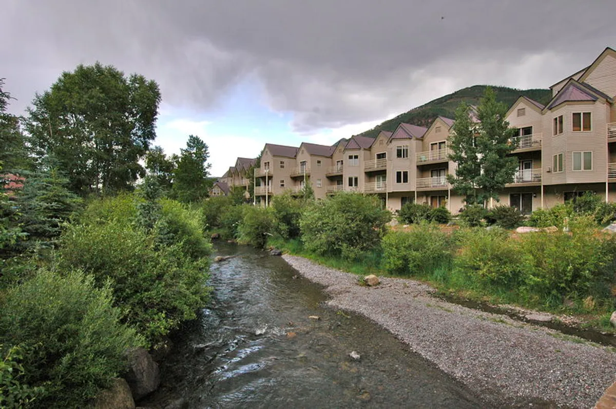 A peaceful river flows alongside lush greenery, with condominium buildings nestled in the background. The stream is bordered by trees and shrubs, offering a tranquil, nature-rich setting perfect for walks or quiet relaxation by the water.