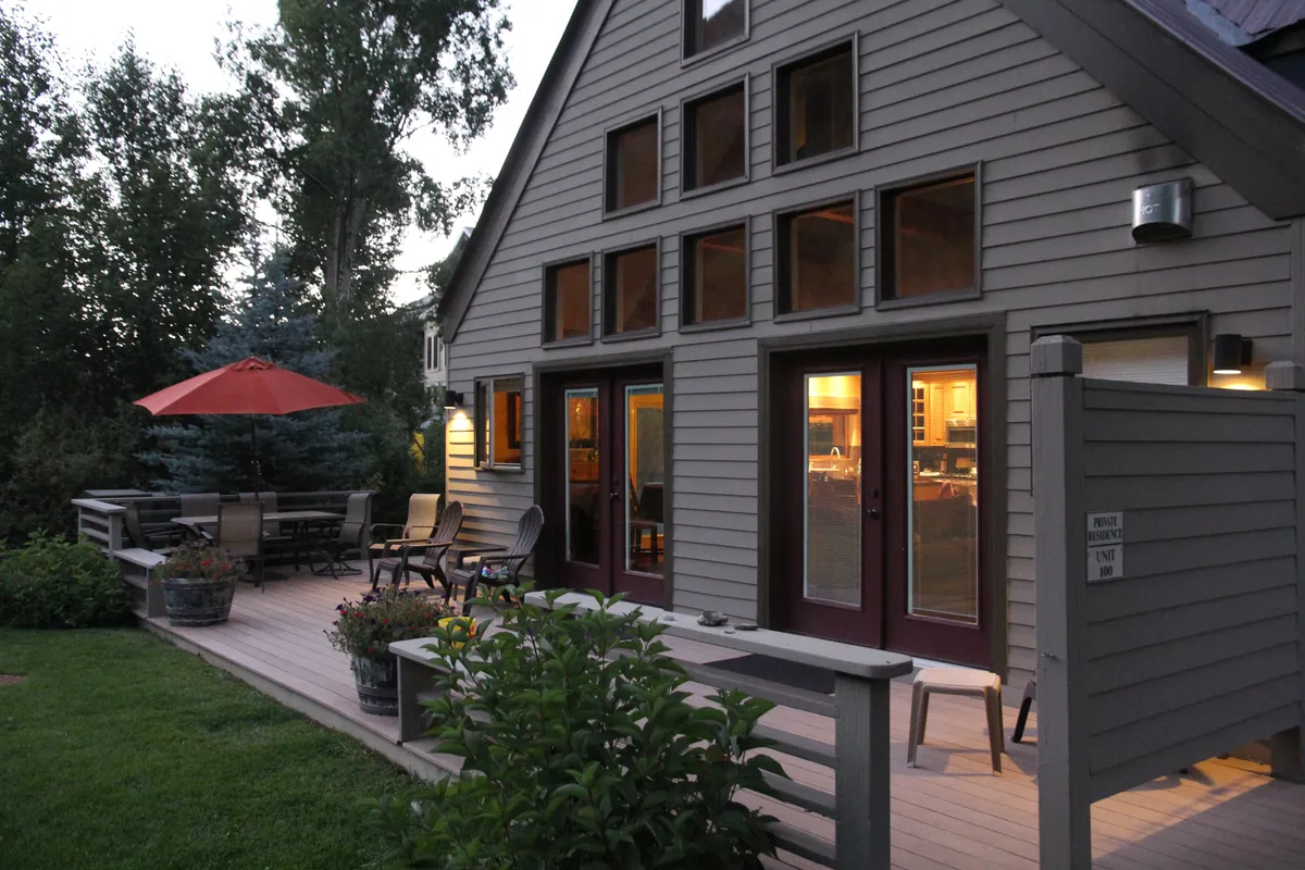 A spacious deck featuring comfortable seating under a vibrant red umbrella, surrounded by lush flower pots and large windows. The scene is set for an evening gathering, with ample space for groups to socialize and relax while enjoying the outdoor ambiance.