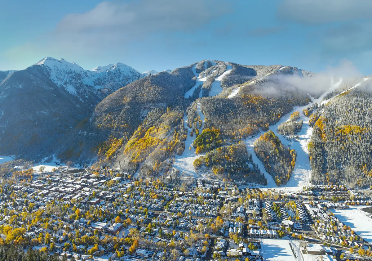 A sweeping view of the town nestled in a valley, surrounded by autumn foliage, snow-capped mountains, and ski slopes. The layout of the town is visible, offering a glimpse of both nature and local amenities.
