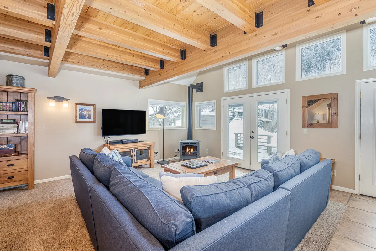 The image showcases a spacious, light-filled living area with a large sectional sofa around a wooden coffee table, a cozy fireplace, and expansive windows that frame breathtaking views. The scene is perfect for relaxing, entertaining, or simply soaking in the mountain ambiance.