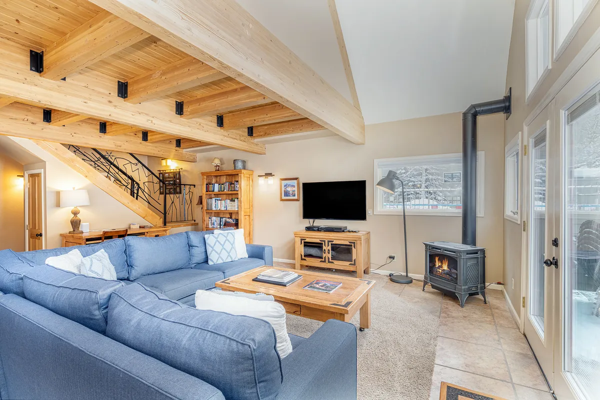 This image features a spacious and inviting living area centered around a large blue sectional sofa and a wooden coffee table. Prominent wooden beams on the ceiling and a stove add rustic warmth, while soft natural light and comfortable seating suggest a place for group relaxation and socializing. A flat-screen TV is visible, enhancing the sense of a communal hub for entertainment and unwinding.