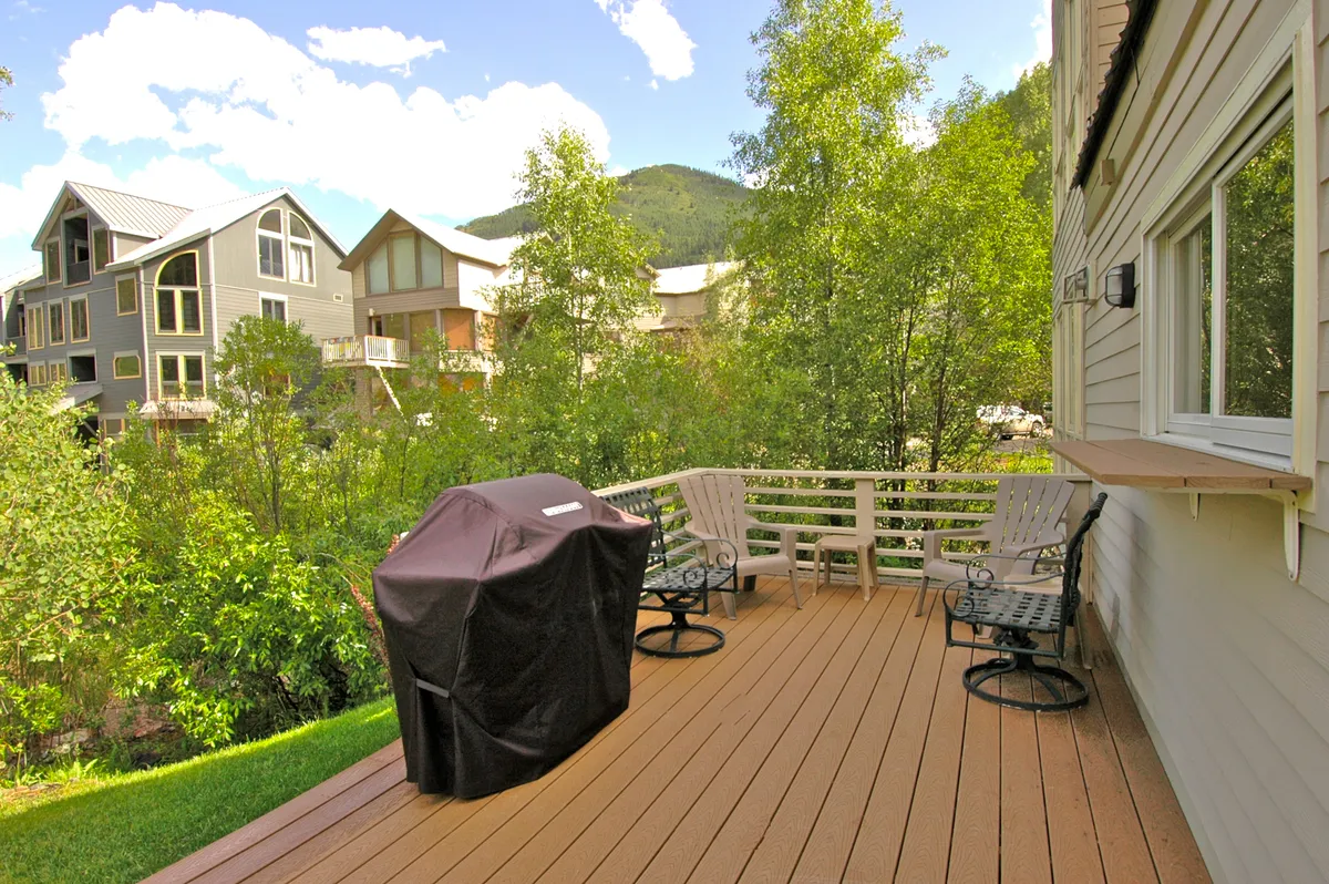 A spacious deck overlooking a panoramic mountain view, with a gas grill prominently featured and surrounded by comfortable outdoor seating. Lush trees frame the scene, and the sun casts a golden glow over the inviting space.