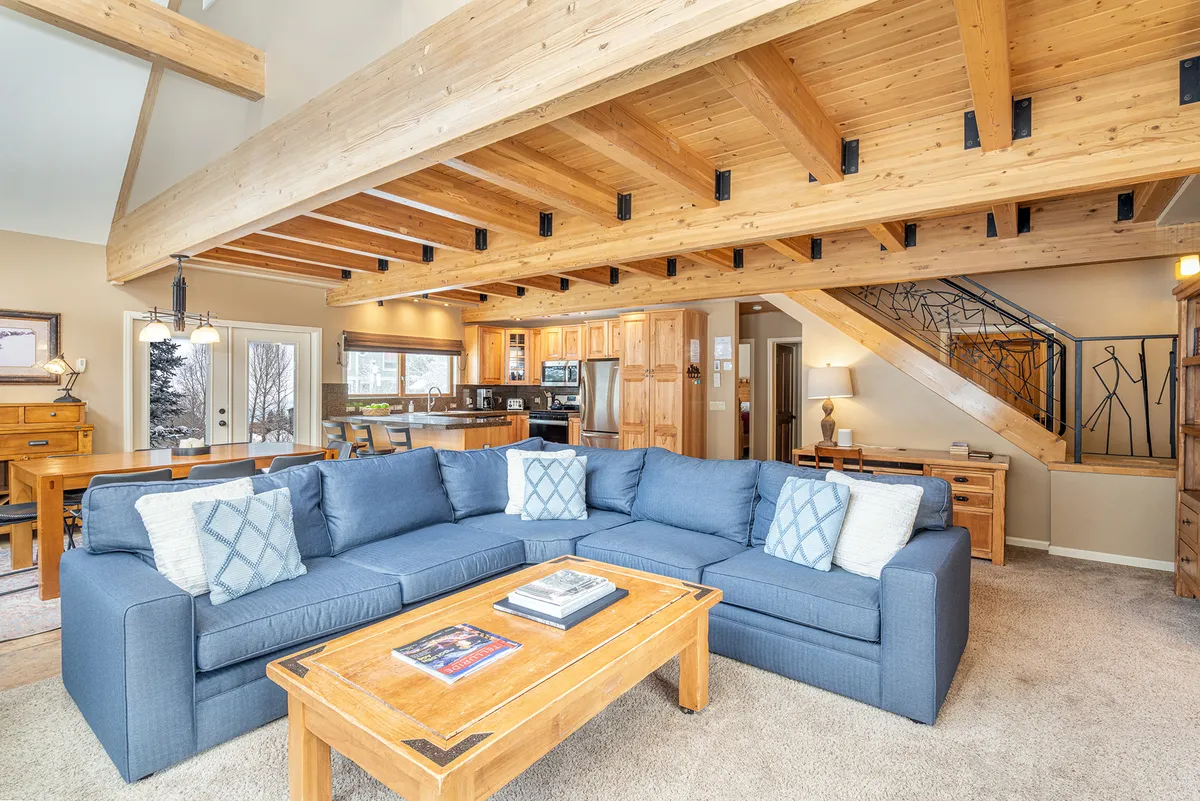 A welcoming open-concept space where a cozy sofa and coffee table blend seamlessly into a visible, fully equipped kitchen. Wooden beams overhead and large windows contribute to a sense of warmth and spaciousness, while the arrangement invites socializing, relaxing, and shared moments.