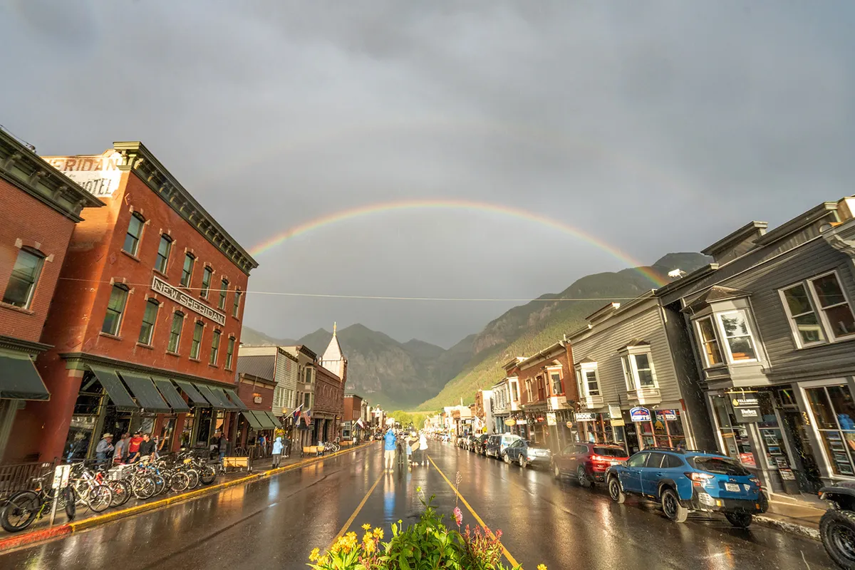 This lively street scene captures Telluride’s historic downtown with its charming buildings, people strolling and biking, and a vibrant rainbow arching above wet pavement. The image conveys bustling local life and the spirit of exploration, with visible shops, outdoor dining, and an inviting atmosphere that draws visitors out to enjoy the town’s offerings.
