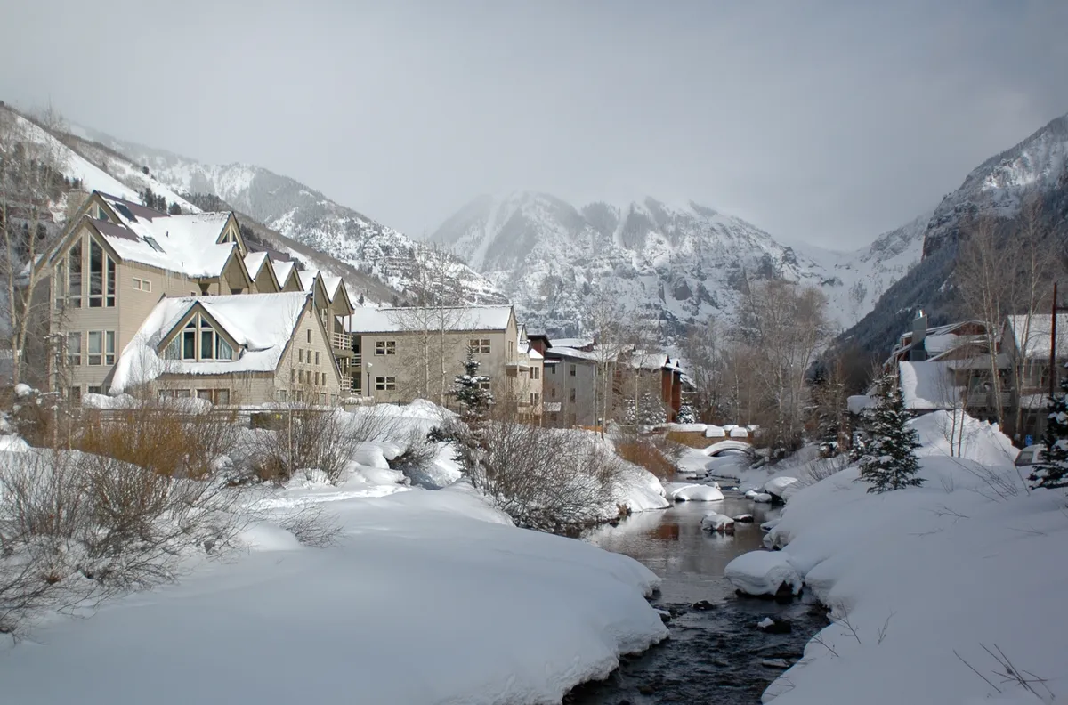 Snow-capped mountains rise above a valley with a river flowing through it, while picturesque buildings dot the landscape. The scene conveys winter tranquility and provides a sense of proximity to skiing and winter outdoor activities.