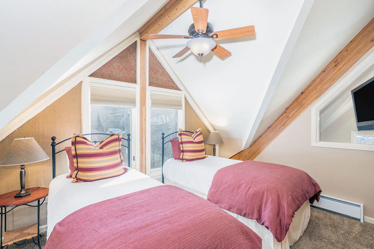A cozy bedroom with two twin beds featuring striped pillows, a window letting in plenty of natural light, and a ceiling fan above. The room exudes warmth and flexibility, perfect for siblings or friends sharing a space.