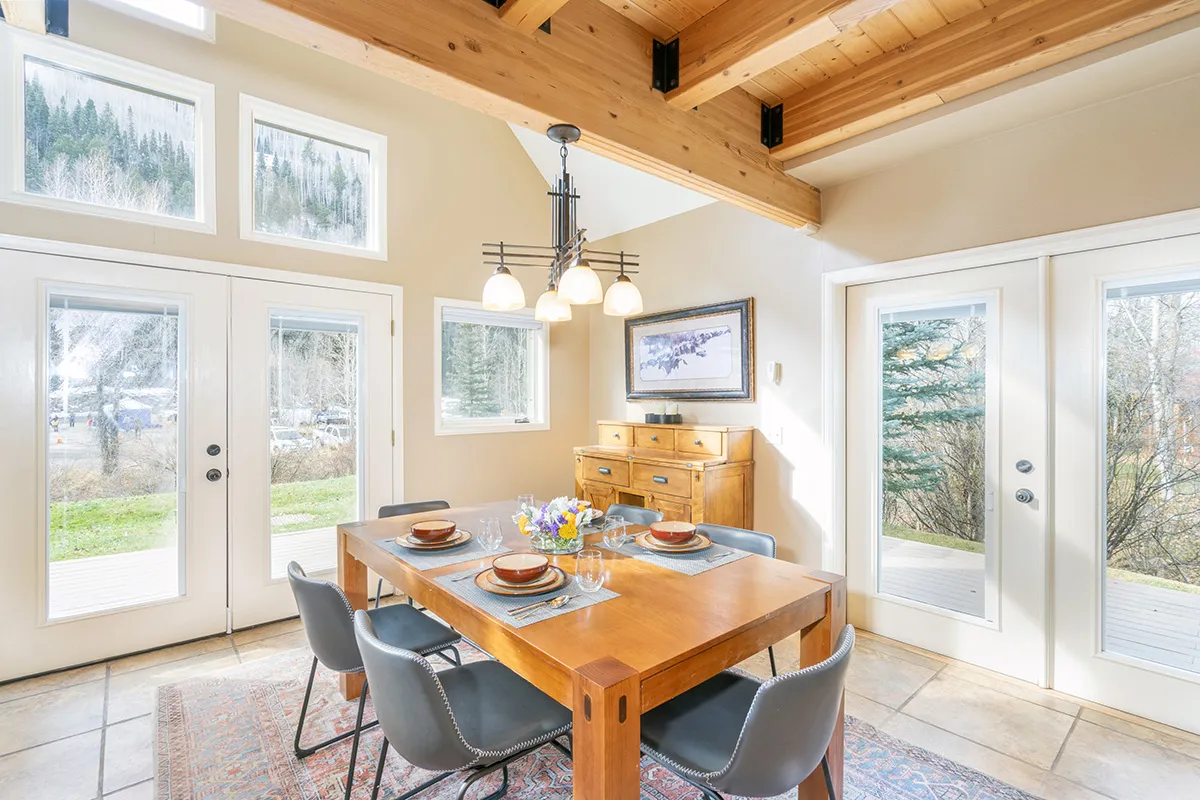 A large wooden dining table sits in front of expansive windows, with wooden beams overhead and tasteful artwork on the wall. The table is set to accommodate a group, perfect for shared meals, celebrations, or planning the next day's activities. Warm lighting and the welcoming setup create a space ideal for memorable group feasts.