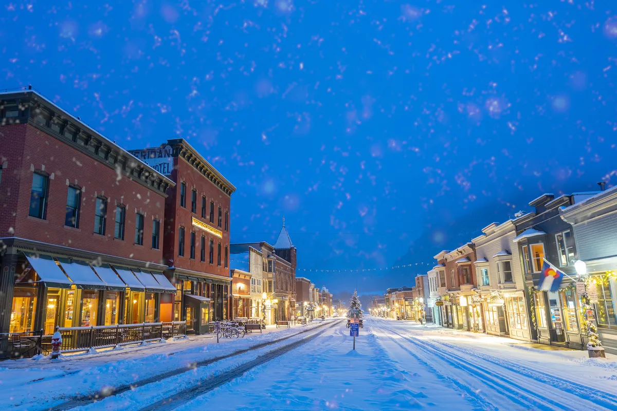 A snow-dusted street lined with historic buildings and festive holiday decorations, including a prominent Christmas tree, captures Telluride’s small-town charm during the winter season. The inviting glow of storefronts and community spirit are vivid, suggesting both shopping and seasonal events within easy reach.