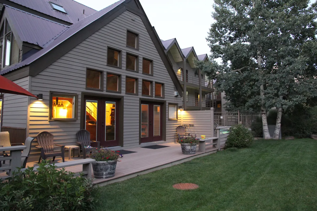 A cozy exterior scene with a beautifully landscaped garden, large windows looking into the home, comfortable outdoor seating, and decorative planters. Subtle string lights set a magical evening mood, and the space is clearly designed for groups to gather and enjoy time together under the stars.