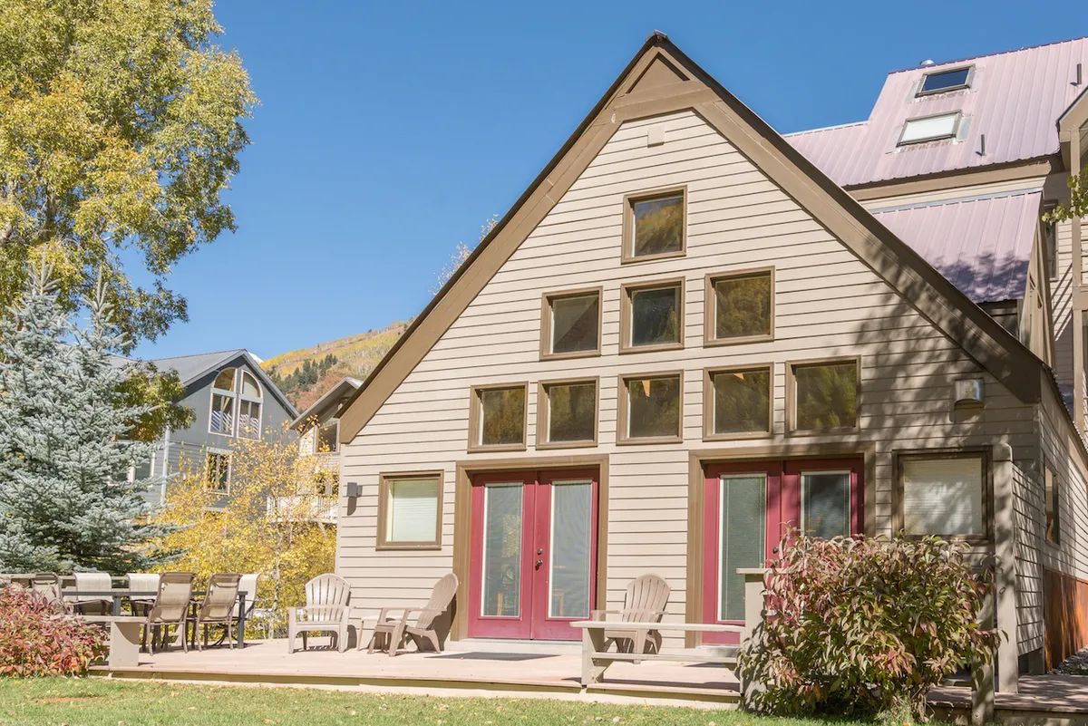 This image features the property's exterior with large front windows, red double doors, and an inviting outdoor seating area. The patio space is set up for relaxing, dining, or socializing, with sunlight illuminating the front of the home. The scene communicates openness and a welcoming atmosphere for families arriving together.