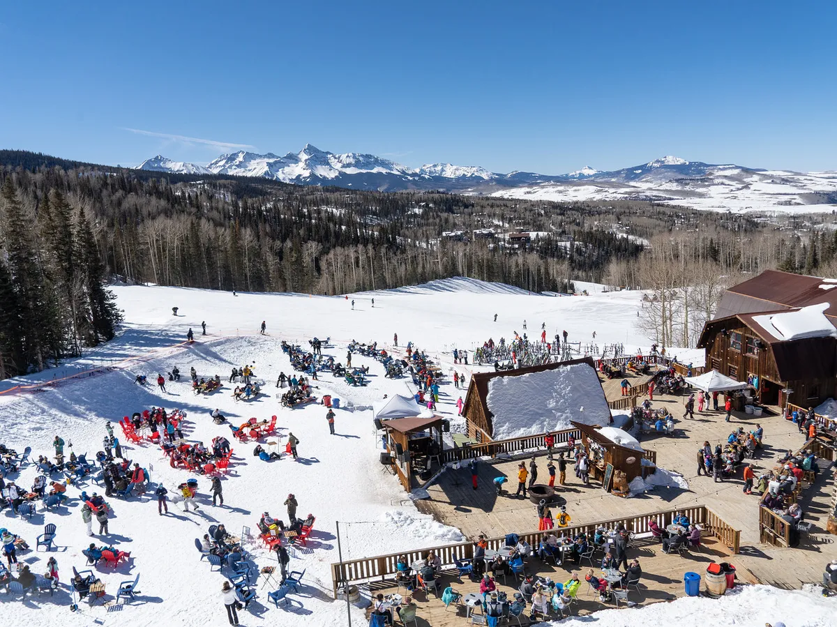 Skiers descend snowy slopes with a lively crowd enjoying outdoor seating near ski lodge buildings. The mountain range provides a dramatic backdrop, and the atmosphere is energetic and social.