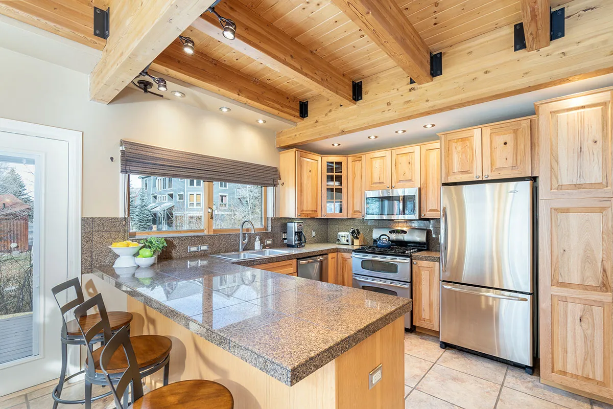 A modern gourmet kitchen with a prominent granite-topped island, stainless steel appliances, and exposed wooden beams. The kitchen is open to the living space, inviting guests to cook, entertain, and gather around. Natural light illuminates the space, enhancing the appeal of the upgraded finishes and practical features.