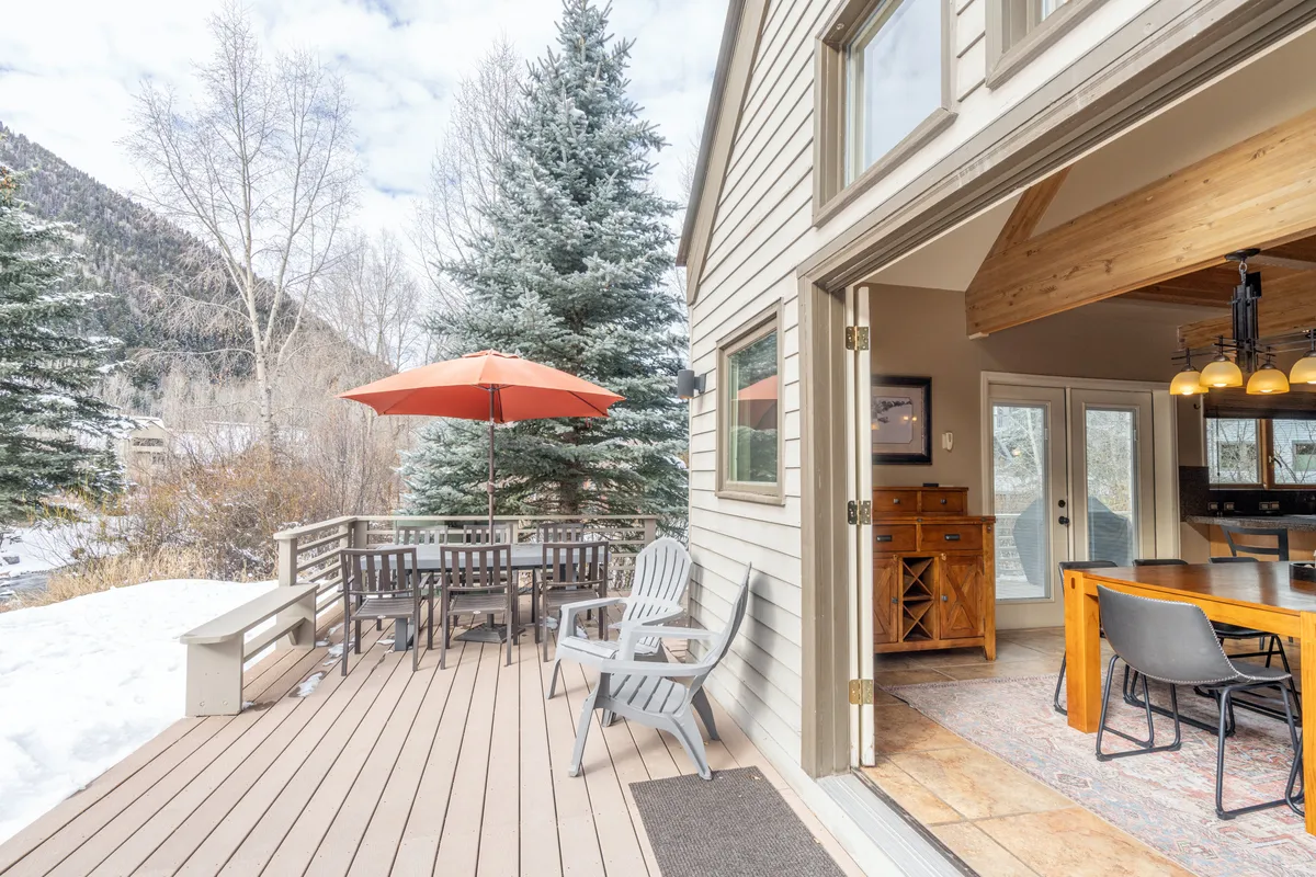 An exterior view showcasing an outdoor dining set beneath a bright orange umbrella, framed by pine trees and large windows. The table is ideal for shared meals, and the scene conveys a relaxed, communal vibe with room for multiple guests.