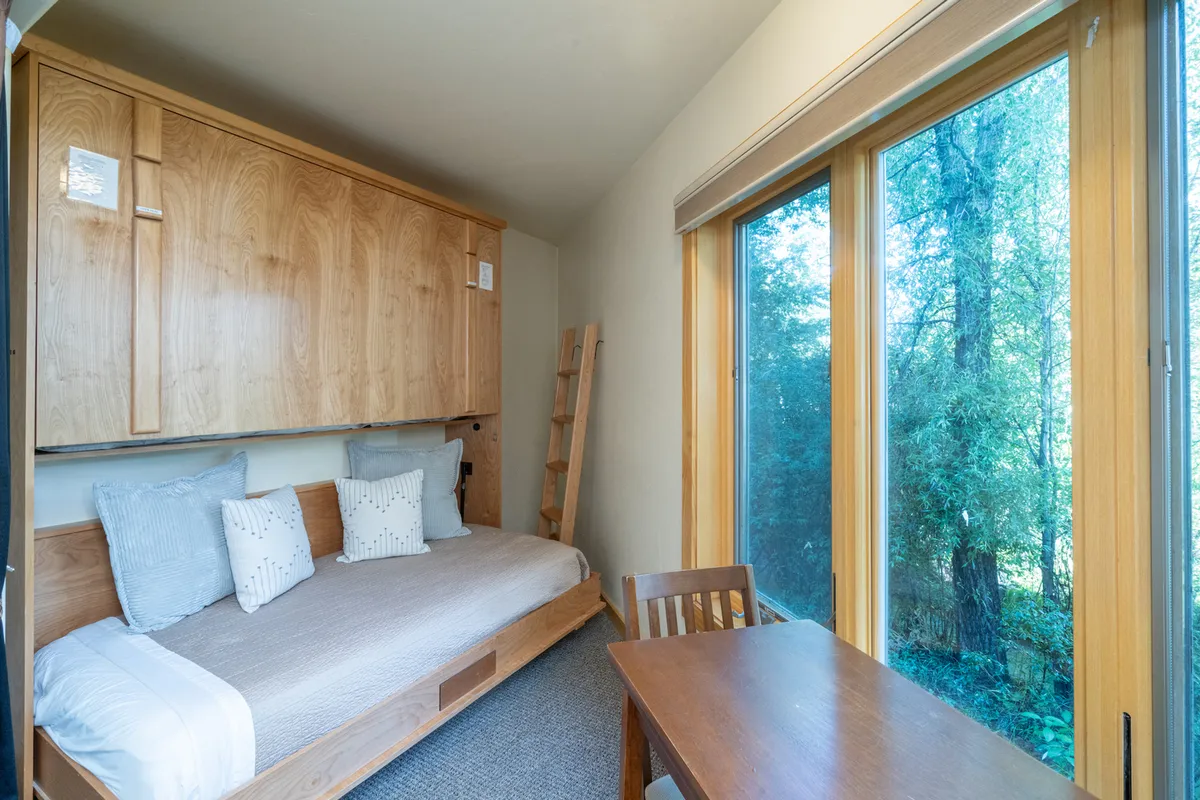 A bright, self-contained studio suite featuring a large bed with cushions, wooden cabinetry, a small table, and expansive windows delivering plenty of daylight. The layout suggests both comfort and self-sufficiency, with space for relaxing, reading, or enjoying a snack.