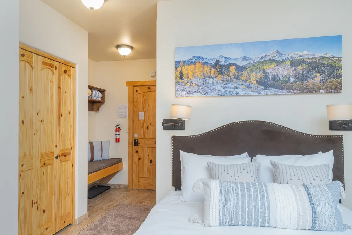 A tranquil bedroom featuring a comfortable bed adorned with decorative pillows, landscape artwork above the headboard, and wooden doors leading to a private space. Natural light filters in through the windows, highlighting the soft linens and giving the room an inviting atmosphere.