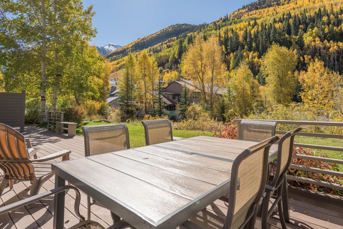 A dining table is set on a deck nestled among trees, with sweeping mountain views in the background. The space is perfect for morning coffee, family meals, or simply soaking up the stunning natural scenery.
